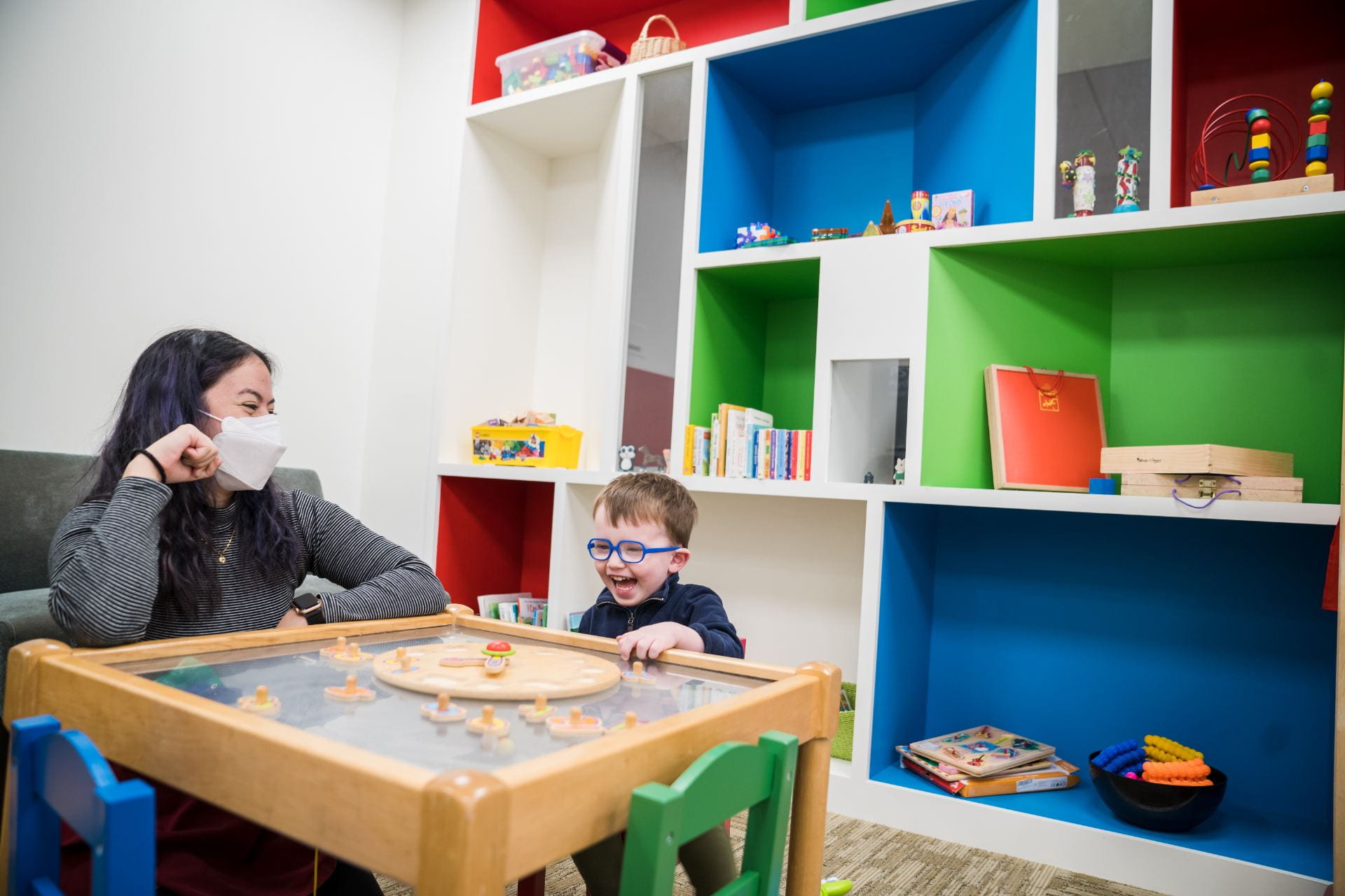 Small child with blue glasses is laughing and playing with a puzzle. An experimenter is playing with them. They are in a colorful lab waiting room.